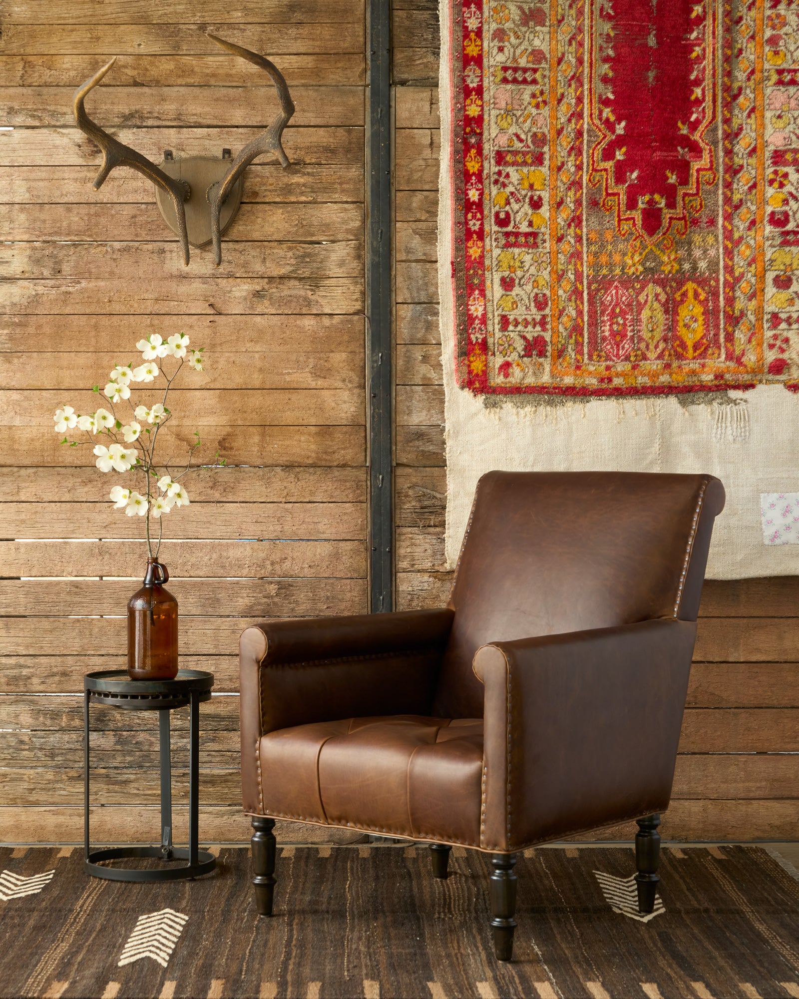  Brown leather chair in front of a wood wall with a tapistry and antlers. A side table has a brown bottle with white flowers. Photographed in Spur Chocolate. 