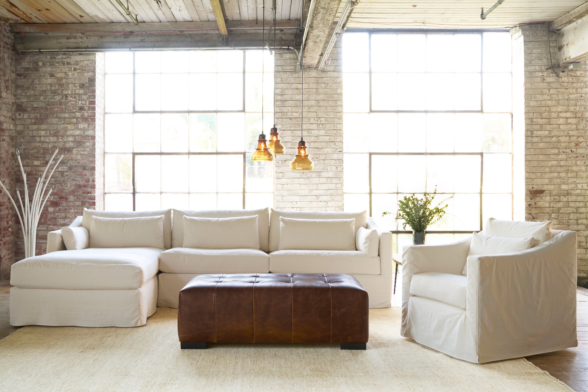  White sectional in front of 2 big windows and next to a white slipcovered chair. Brown leather ottoman in the middle and 3 brown glass pendants. Photographed in Molino Ivory. 