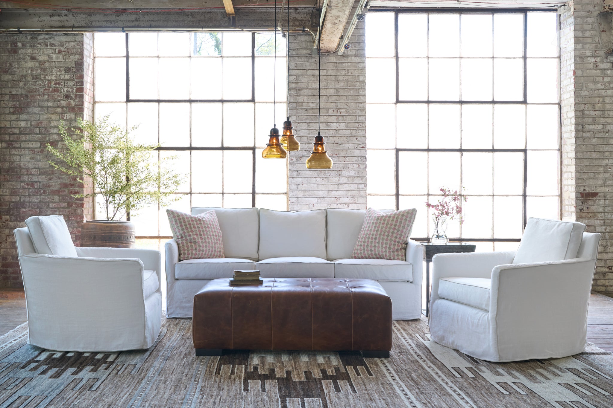  White sofa in a showroom with 3 glass light pendants, 2 white chairs on the side and a brown ottoman in front. Photographed in Ashville White. 