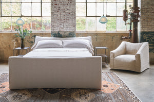  Bed in a showroom with large windows. A slipcovered chair is on the right side. Two glass pendants are hanging on each side. Photographed in Adler Sand. 