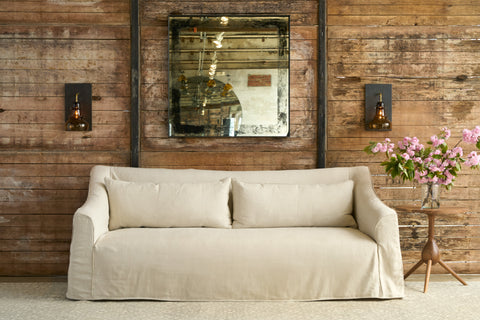 Slipcover sofa in front of a wood wall. Mirror on the wall with 2 sconces. Side table with pink flowers on top. Photographed in Noah Bone.