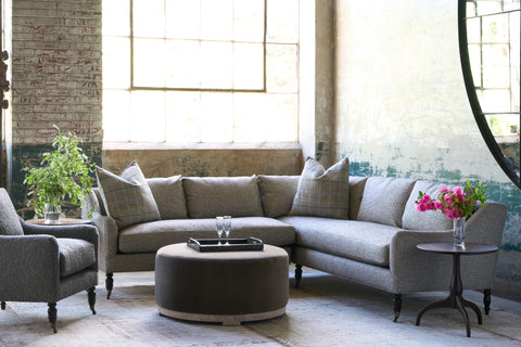 Sectional sofa in front of a large window with an ottoman in front. Tray on top with 2 glasses. Chair on the left and wood side table on the right with pink flowers on top. Photographed in Ferrara Charcoal.