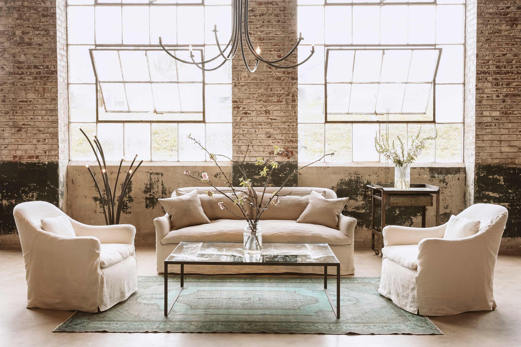  Daytime in a showroom with large windows and brick wall. The Hazel sofa is slipcovered in Quixote Oatmeal, with 2 Hazel Chairs on each side, also slipcovered in a white linen. The coffee table is rectangular with a mirror top. There is a floor lamp with multiple branches on the left and a Ramo chandelier hanging from the ceiling. Photographed in Brevard Burlap. 
