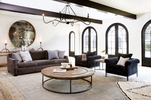 Grand living room, daytime, the Hayden sofa is upholstered in Matteo Dark Grey. There are 2 Henrietta Chairs in a black velvet and a large round wood coffee table. There is a Spider Chandelier hanging from the ceiling. A large Geller mirror is hanging on the left wall. Photographed in Matteo Dark Grey. 