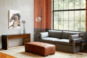  Living room setting. Slipcovered sofa in grey fabric and brown leather ottoman placed in front of brick wall with large window. Wood console is also placed against one wall with art work of a horse and floor lamp placed next to console.  
