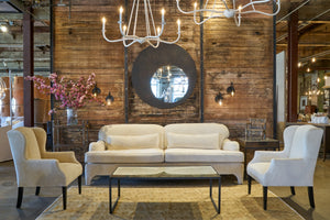  White sofa in front of a wood wall with a mirror. Wing chairs on each side and white chandeliers. Photographed in Velluto Natural. 