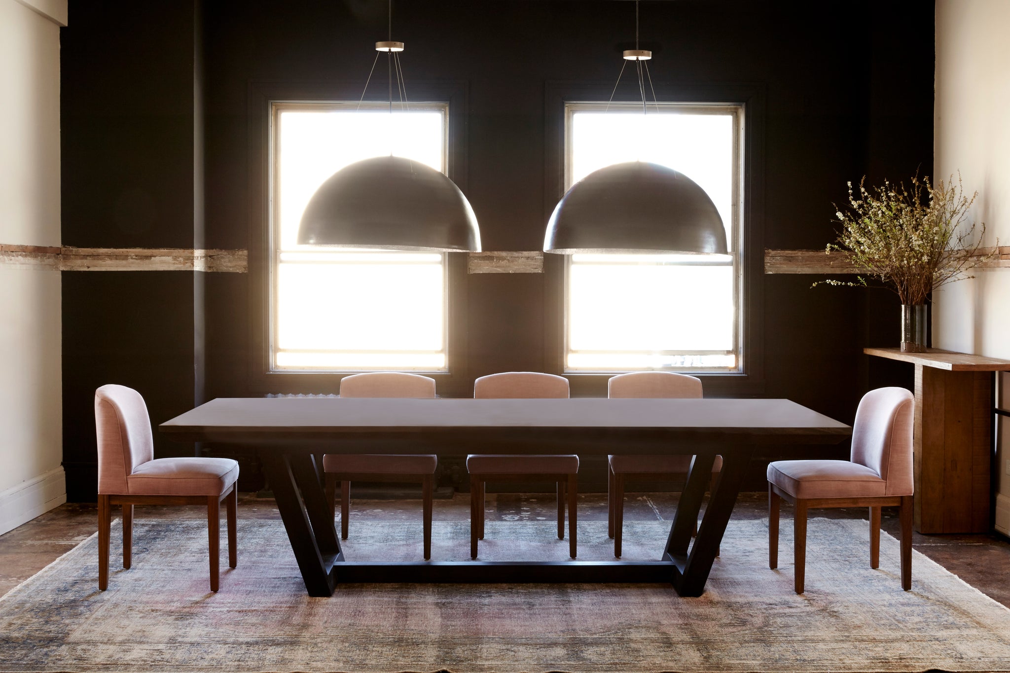  Dining room photo. Rectangular dining table with five dining chairs placed around it which are upholstered in a pink fabric and have wood bases. There are two black circular pendants hanging above the dining table and the room has a black accent wall with two large windows. All the furniture is sitting on top of a grey rug.  