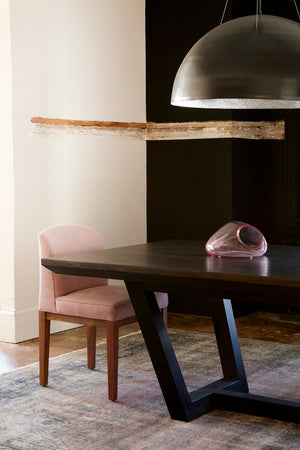  Dining room detail shot. Upholstered dining chair with wood base and pink fabric placed at the end of a dining table in a dark wood finish. Black circular pendant hanging above dining table.  