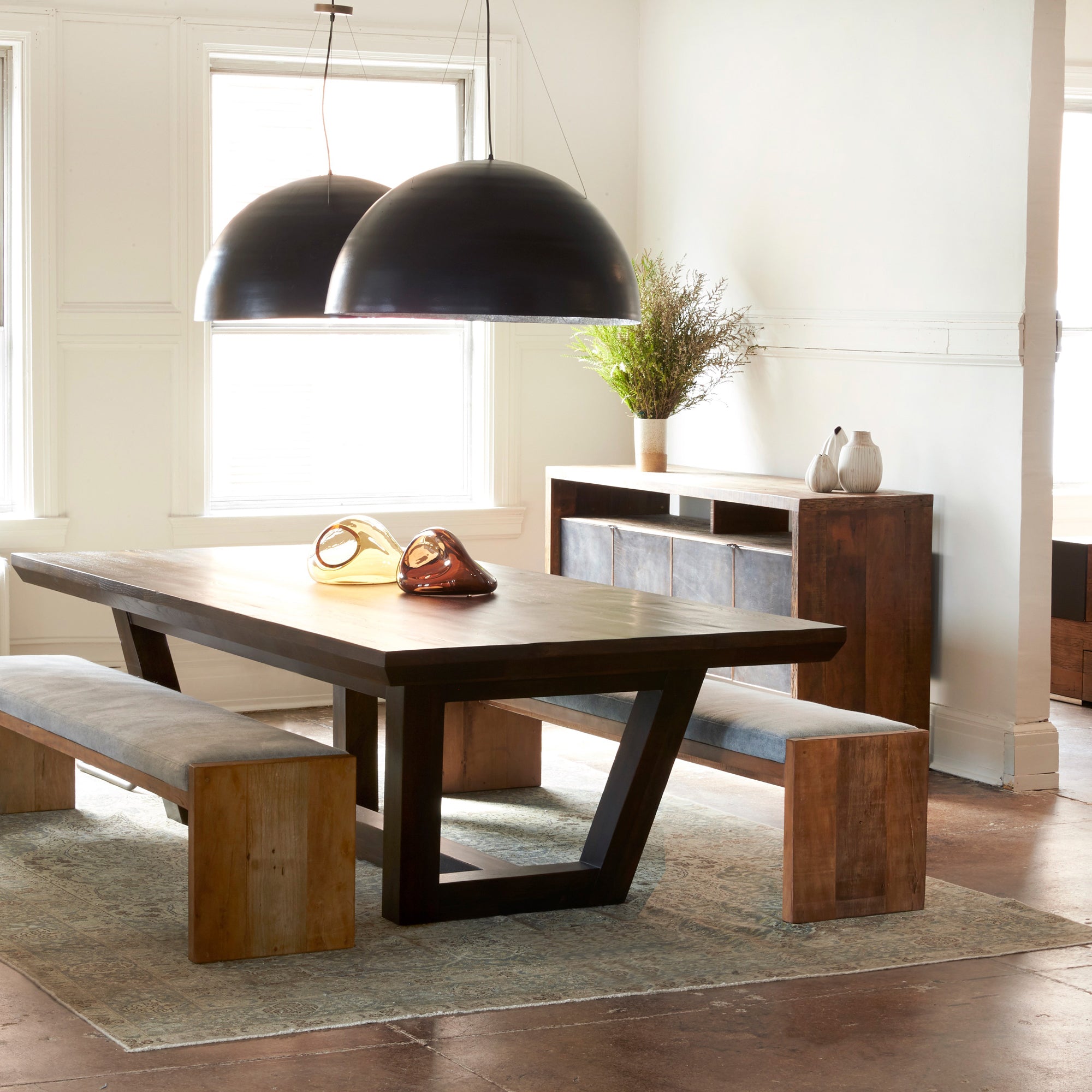  Dining room photo. Rectangular dining table with two upholstered benches on either side as seating. There are two black circular pendants hanging above dining table. The dining set is placed in a white room with a large window, the wall adjacent to the window has a wood credenza against it.  