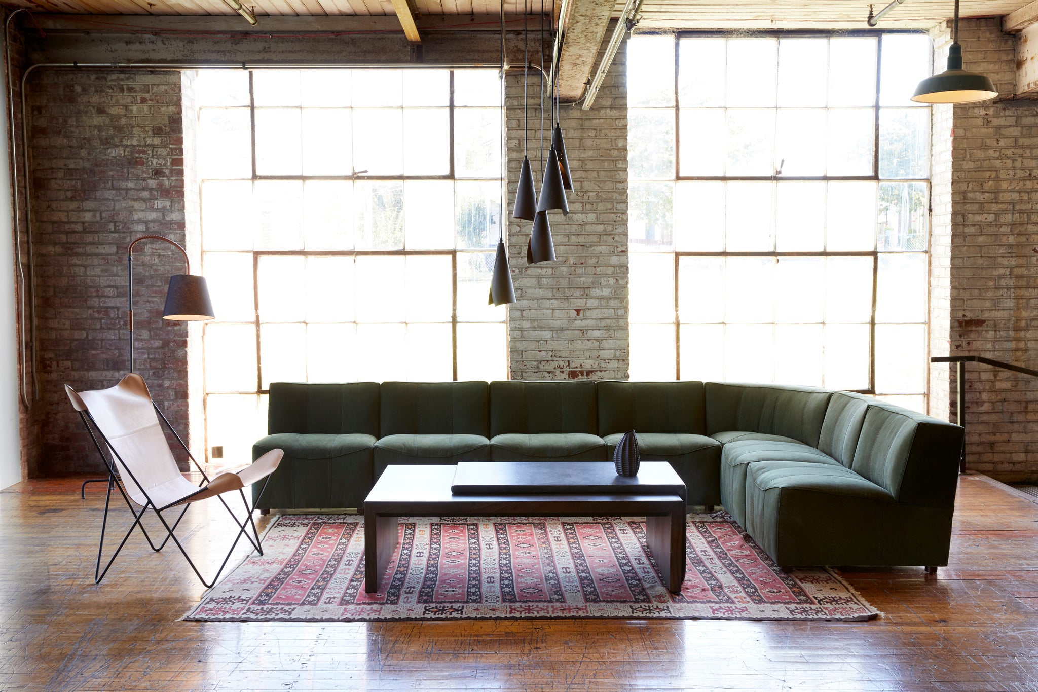  Century Sectional sits in the middle of a room with a wood coffee table and leather chairs. There is also a red patterned rug underneath the pieces. In the background is a brick wall with large windows. Above the pieces hangs multiple lighting lamps. Photographed in Hunter Green. 