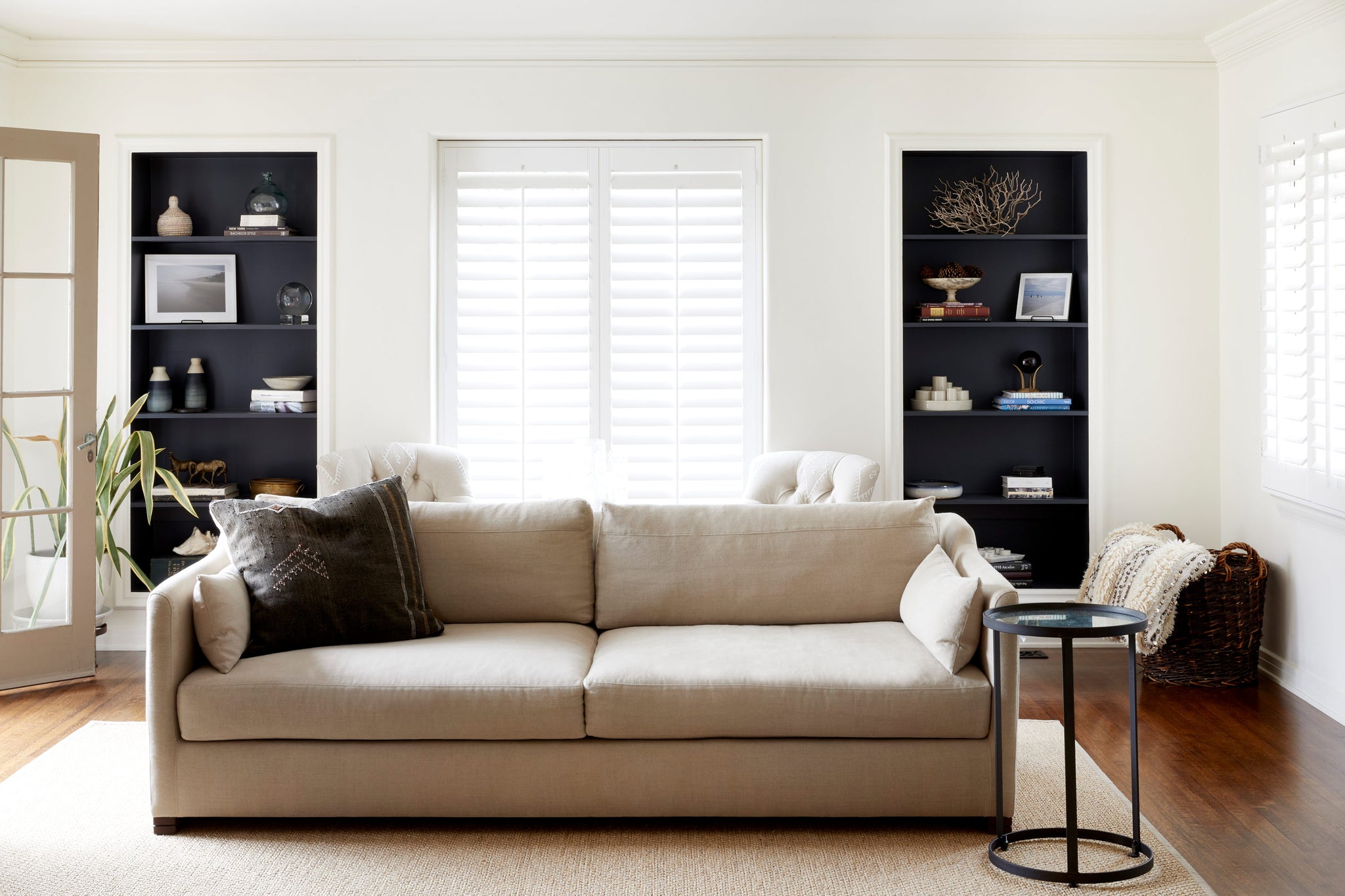  Daylight in a white living room with 2 black bookcases on the back wall. The sofa is in a natural color fabric with a round side table to the right. Photographed in Brevard Burlap 