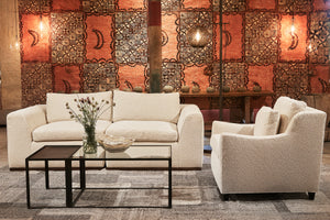  The 2 piece Lorenzo Sofa in Segura Natural is sitting with the Milo Chair in the same fabric in front of a bright colored Tapa cloth in the background. There is a 2 piece Welders coffee table in the middle with flowers and a bowl on top. There is an Aurora floor lamp in the back. Photographed in Segura Natural. 
