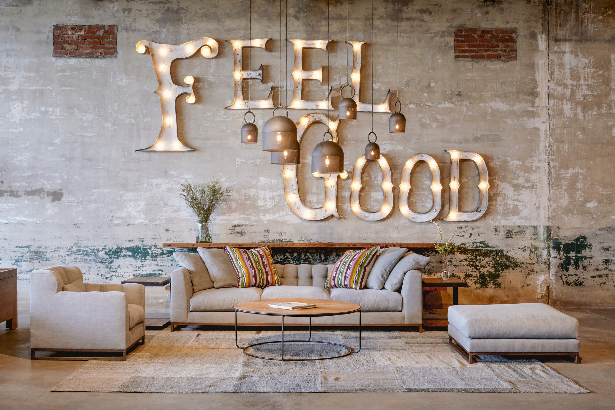  Upholstered sofa with matching chair and ottoman in light grey fabric. Round wood coffee table at the center with cluster of oxxo pendants hanging above. Back wall has a metal lit up sign reading "Feel Good".  
