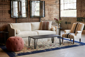  Pouf Ottoman 26 in Rye Terra next to a coffee table, a light sofa, and chair. Photographed in Rye Terra. 
