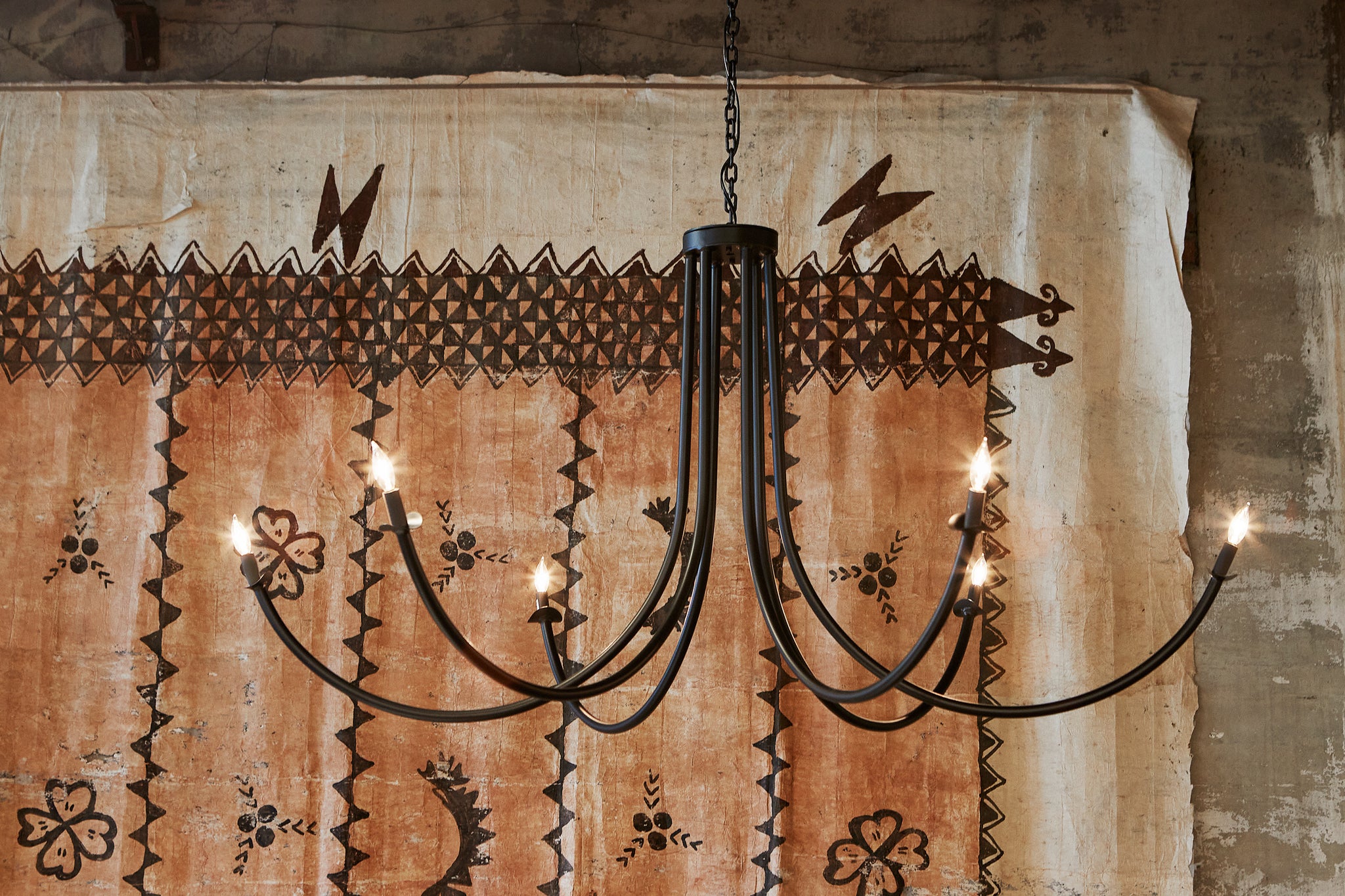  Black metal chandelier with 6 bulbs hanging in front of a brown painting. 