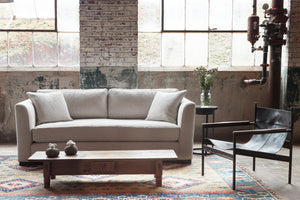  The Ryder sofa is in Bellamy Natural in a bright room with aged brick walls and large industrial windows. A brown leather chair is on the right, a Rotor side table is next to the sofa with flowers in a vase. The low vintage wood coffee table has 2 small flower arrangements on top. There is an industrial pipe in the back right corner. Photographed in Bellamy Natural. 