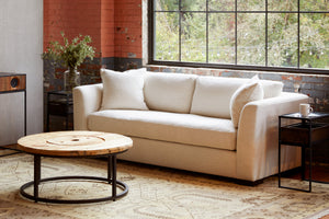  Ryder sofa in Lester Snow in a loft style room. Round coffee table with wood top in front. In the background, large window, and wood and metal side table on each side with a vase with greenery on the left. Photographed in Lester Snow. 