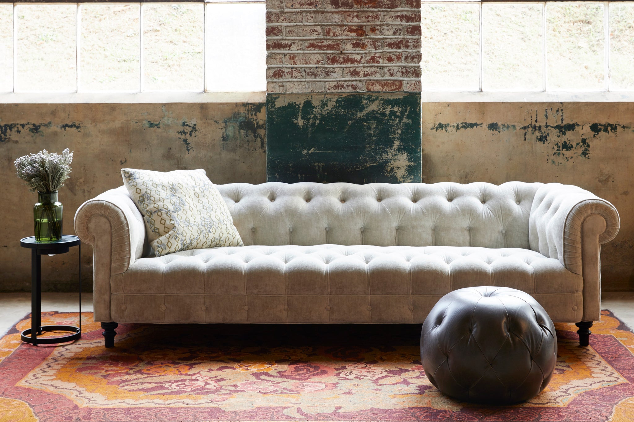  Cream velvet tufted sofa in front of windows with a leather pouf in front. Photographed in JD Velluto Stone. 