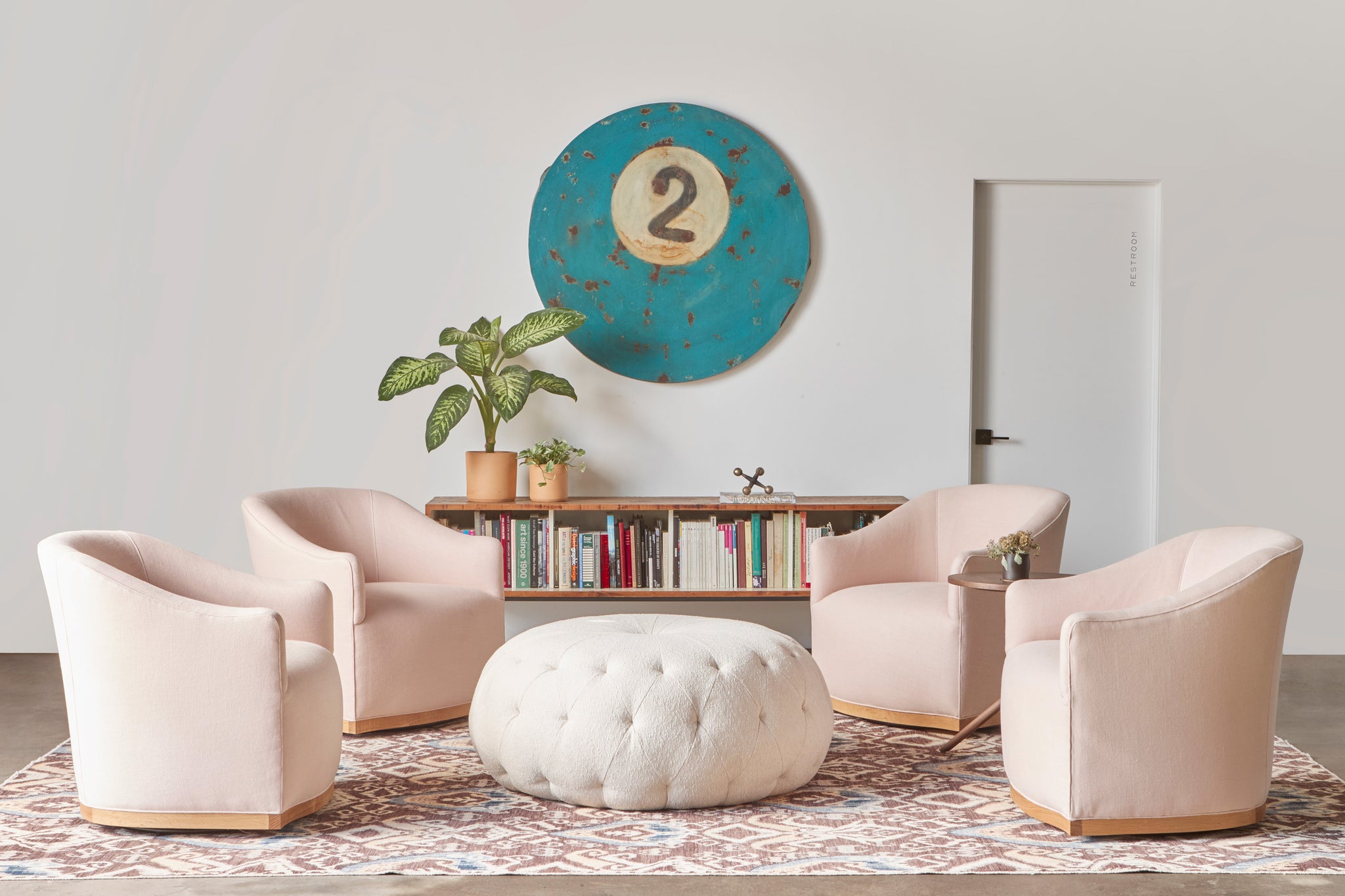  Four pink chairs around a tufted ottoman in an oatmeal fabric. There is a billiard ball artwork in turquoise on the wall above a credenza with books. Photographed in Brevard Rose. 