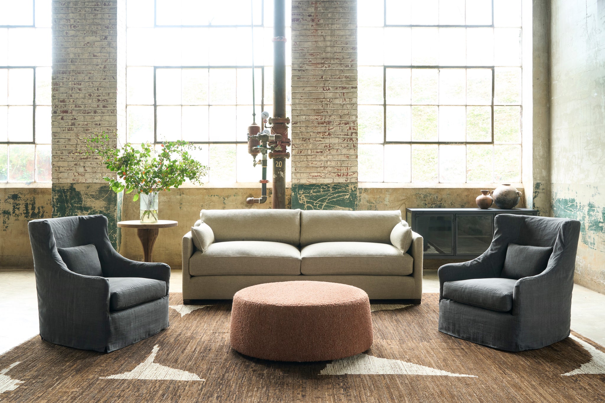  Beige sofa in a showroom with 2 black wing chairs on each side. An ottoman in an orange textured fabric is in the middle. Flowers on a table on the let side. Brown and white rug. Photographed in Knobby Canyon. 