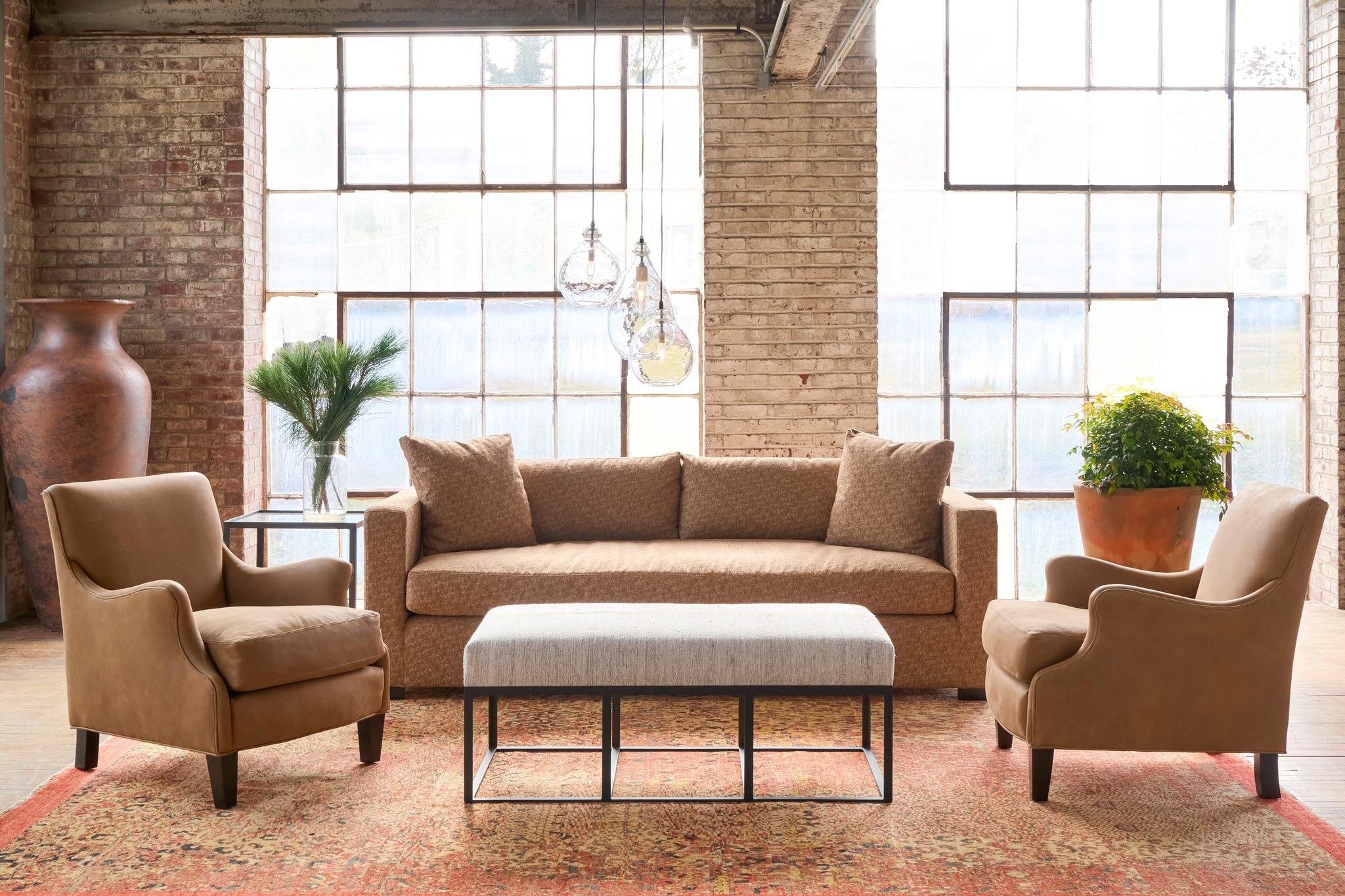  Brown sofa with matching armchairs and a coffee table in a room with large windows and brick walls. Photographed in Lucerne Hazelnut. 