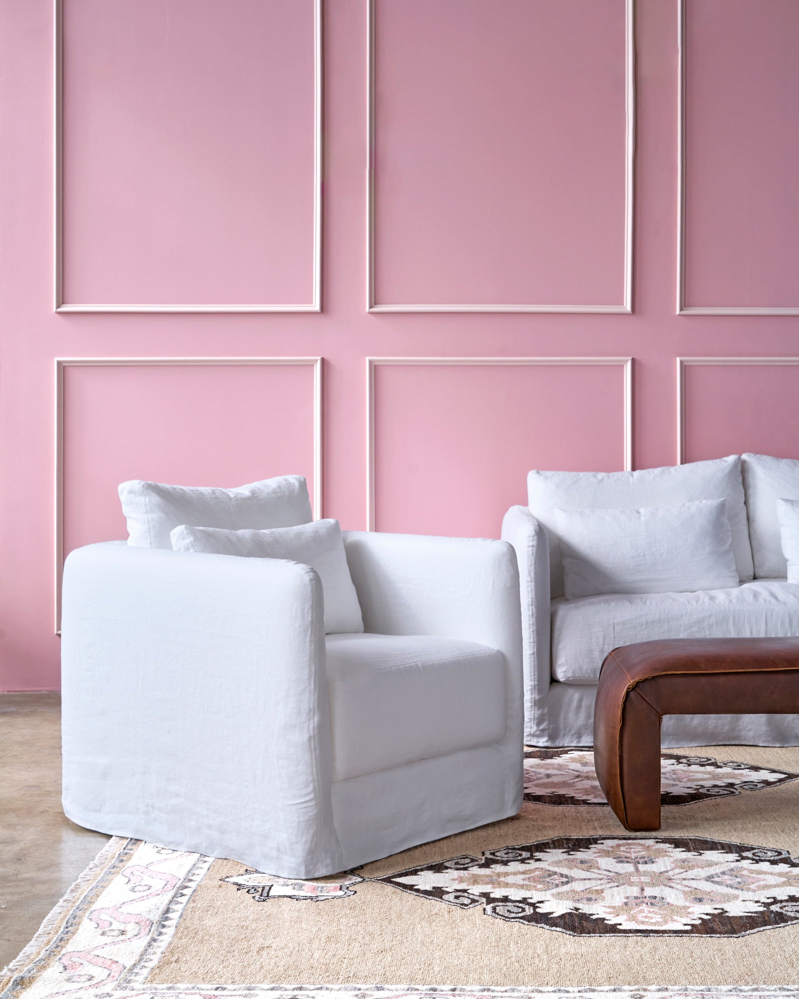  White slipcovered chair in front of a pink wall and  next to a white sofa and leather bench. Photographed in Otis White. 