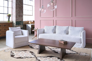  White sofa in front of a pink wall with a rug underneath and a white chair on the left. Walnut coffee table in front. Photographed in Otis White. 