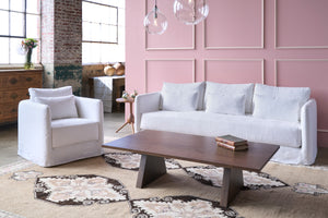  White slipcovered chair in front of a pink wall and  next to a white sofa and leather bench. Photographed in Otis White. 