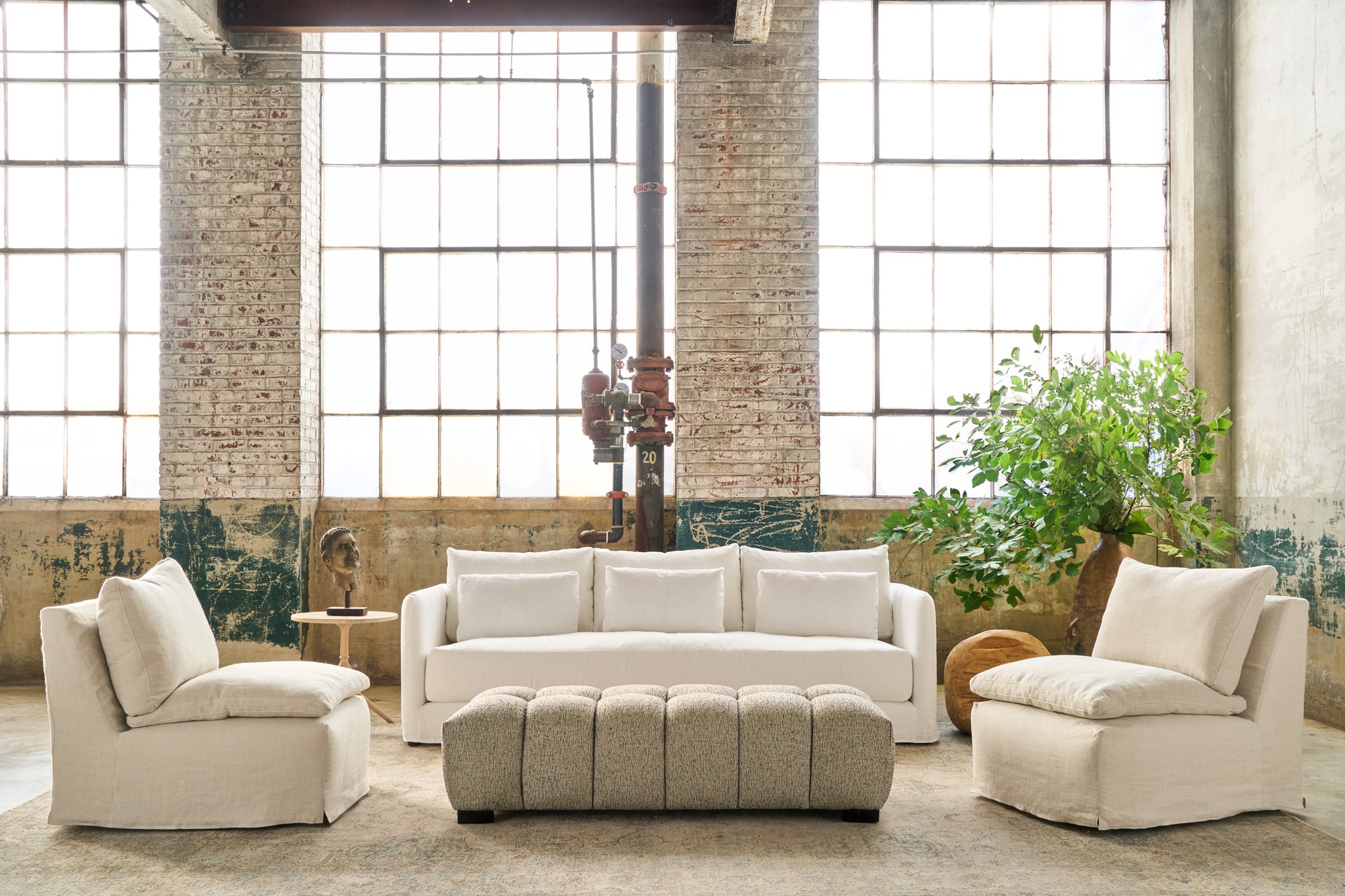  White sofa in a showroom in front of 3 large windows. An armless chair on each side, a tufted ottoman in front. Plants in a pot in the right corner. Photographed in Otis White. 