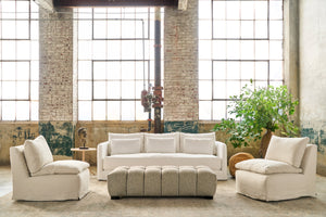  White sofa in a showroom in front of 3 large windows. An armless chair on each side, a tufted ottoman in front. Plants in a pot in the right corner. Photographed in Otis White. 