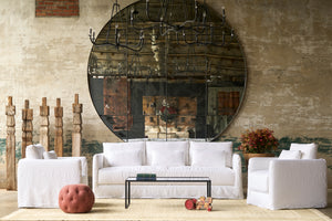  White sofa with 2 white chairs in front of a large round mirro. 5 wood sclptures on the left. A red pouf in front of the left chair. Metal coffee table with vintage toy on the ground. Red plant on the right. Photographed in Otis White. 