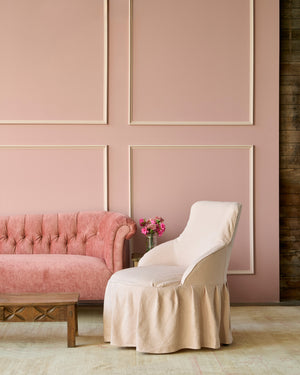  Slipcovered chair in pink with a ruffled skirt. In front of a pink wall and next to a ink velvet sofa. Photographed in Brevard Rose. 