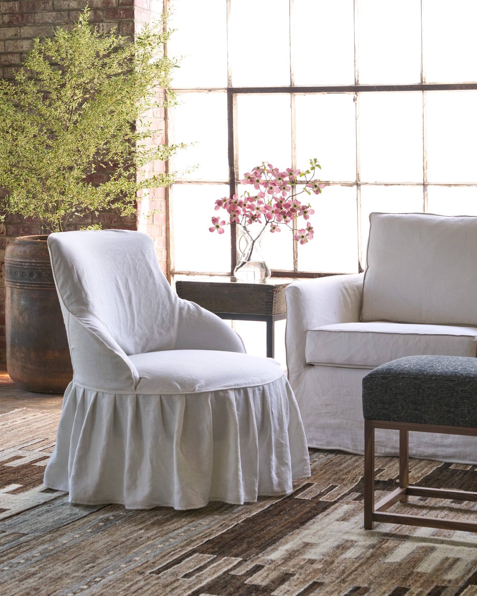  Chair in a showroom next to a white sofa on a brown rug. Side table with pink flowers. Photographed in Otis White 