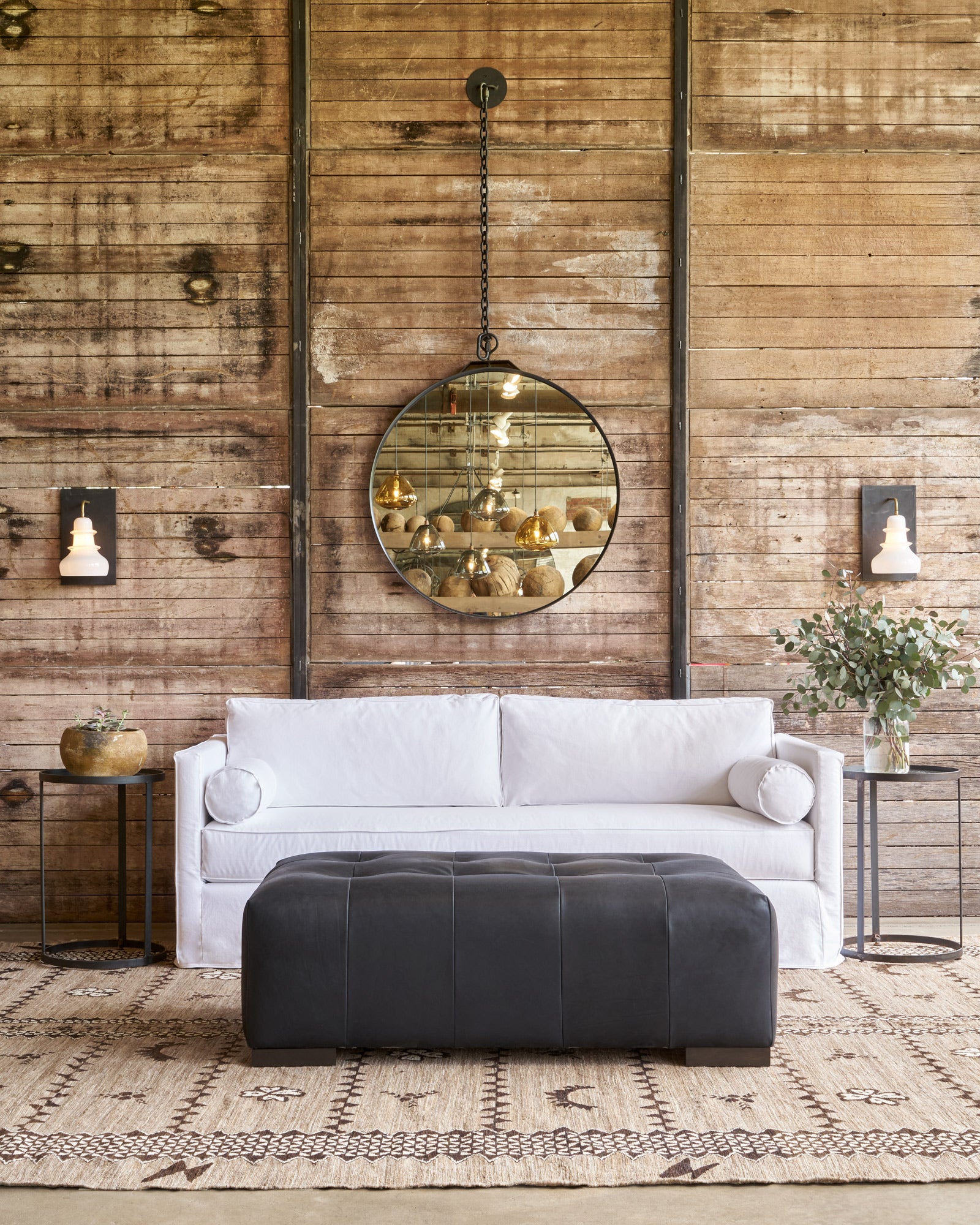  White sofa in front of a wood wall with a round mirror. Black leather bench in front. Photographed in Bracco Black. 