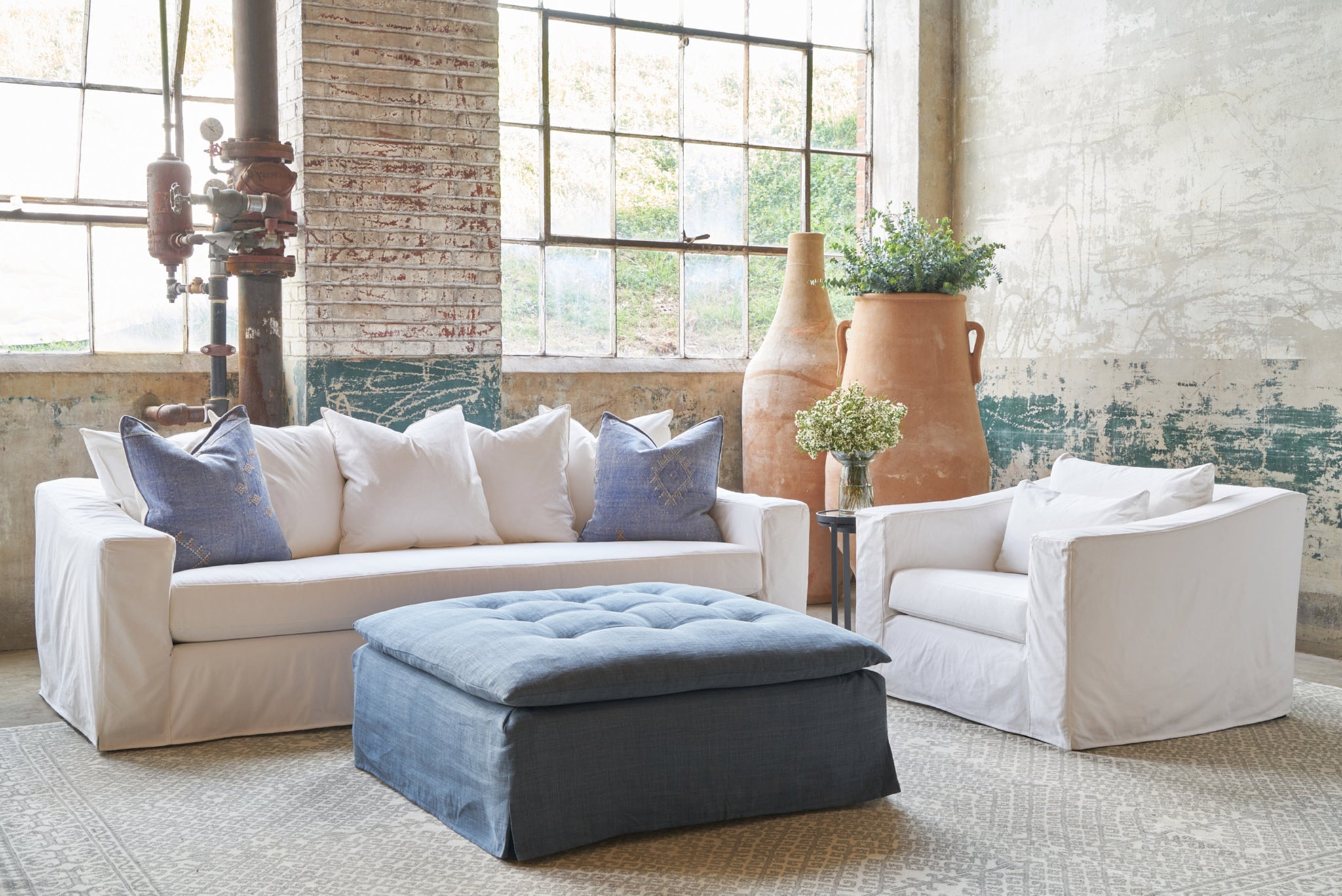  Showroom with large windows, a white sofa with blue accent pillows and a white chair on the right and a blue slipcovered ottoman in the center. Photographed in Luna White. 
