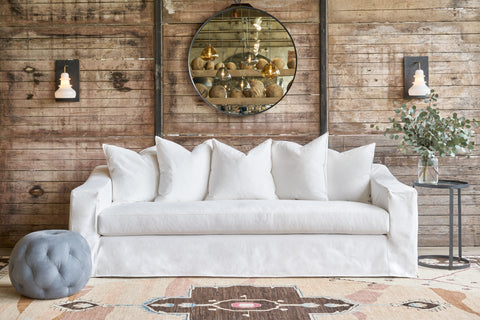 White sofa in front of a wood wall with a grey leather pouf in front. Photographed in Luna White.