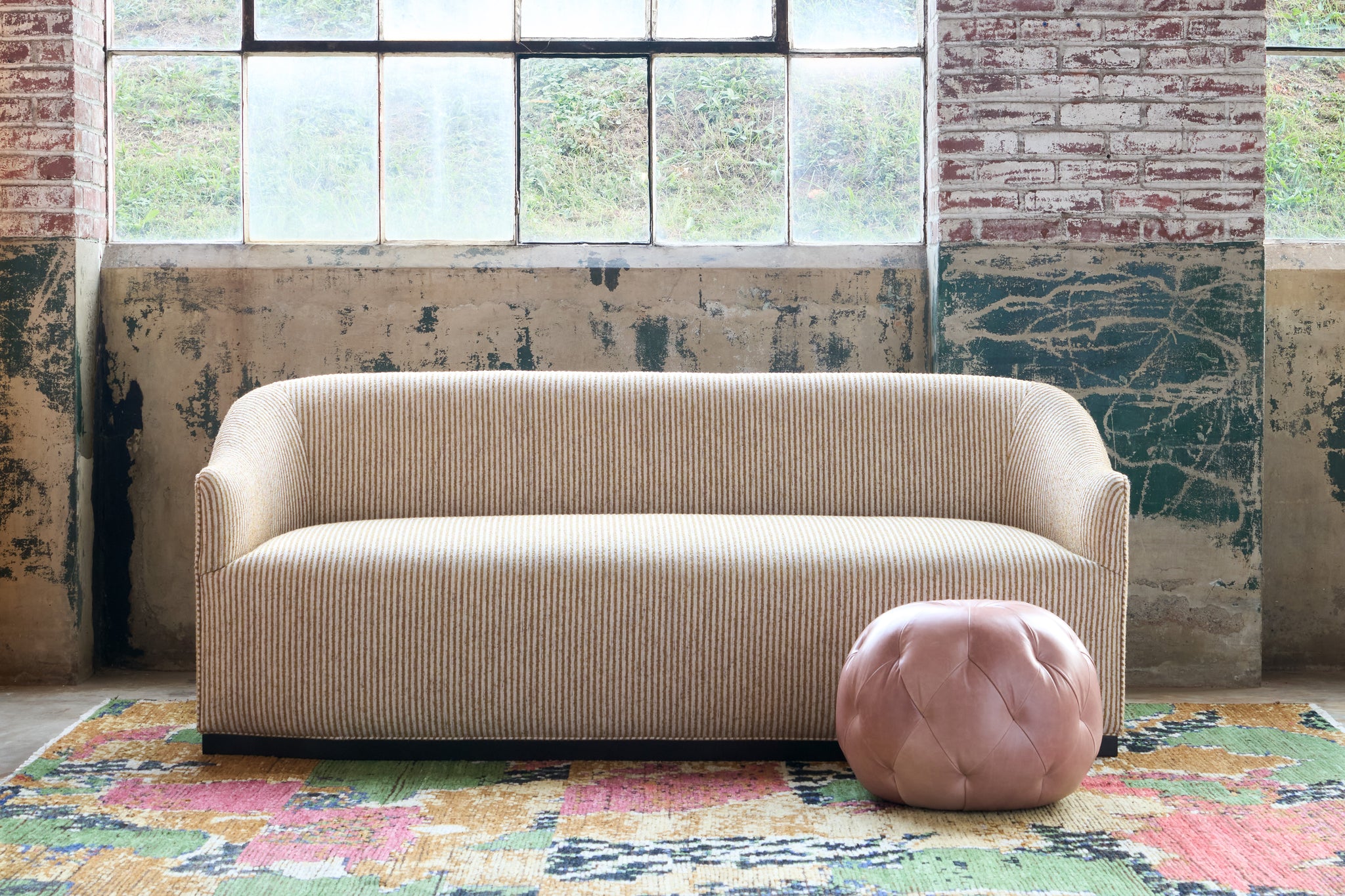  Striped sofa with a pink pouf on a colorful rug against a textured wall. Photographed in Shiloh Turmeric. 