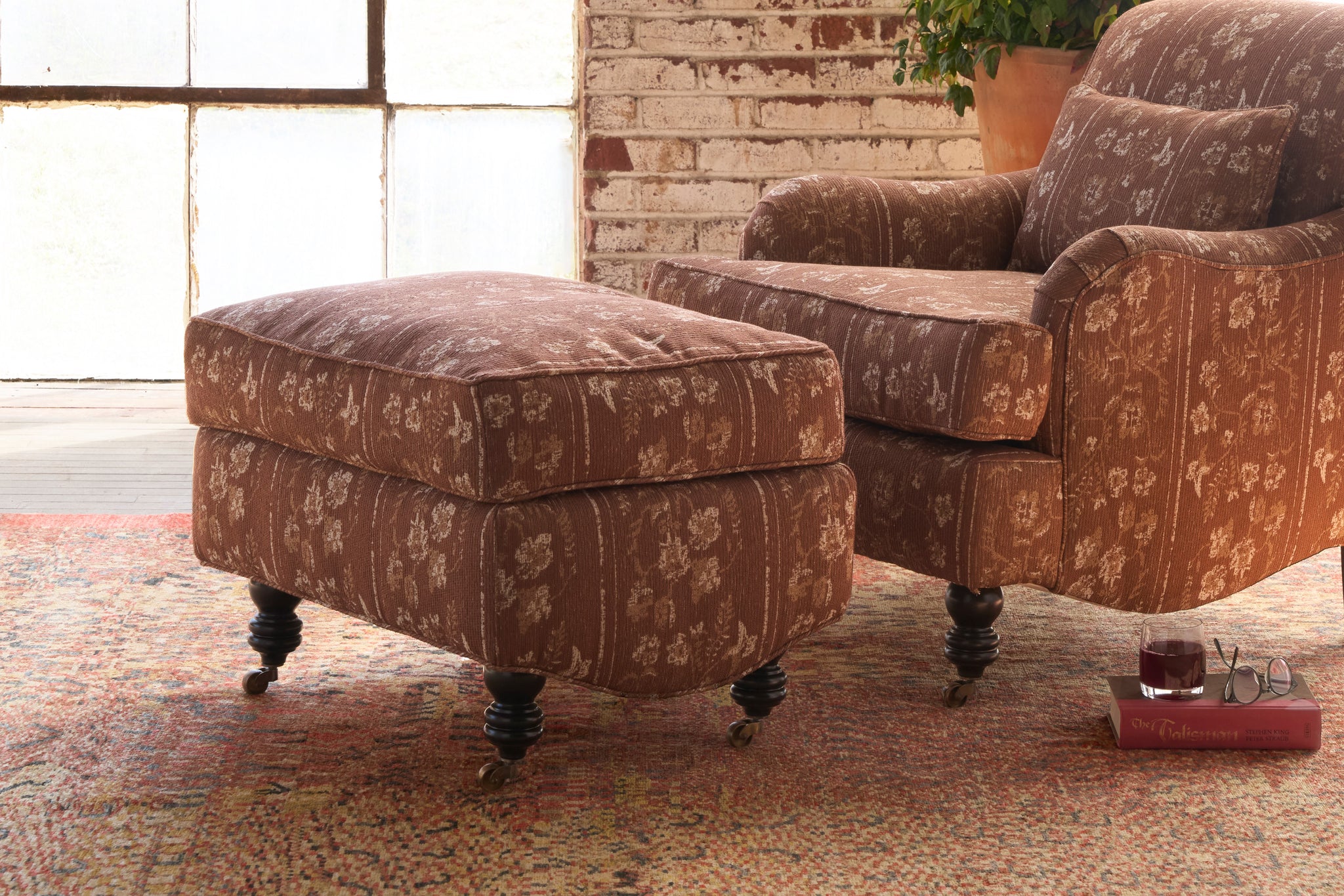  Brown patterned armchair and ottoman on a textured rug with a brick wall background. Photographed in Foret Rust. 