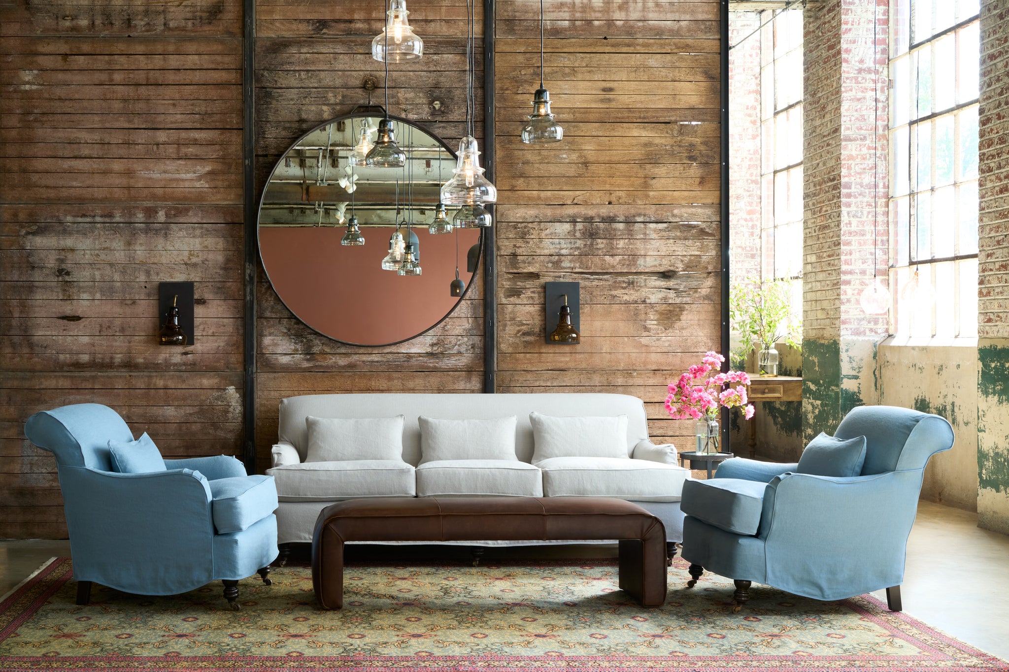 Pale colored sofa and 2 blue chairs in front of a wood wall. Large round mirror on the wall. Glass pendant lights above a brown leather bench. Photographed in Brevard Ciel. 