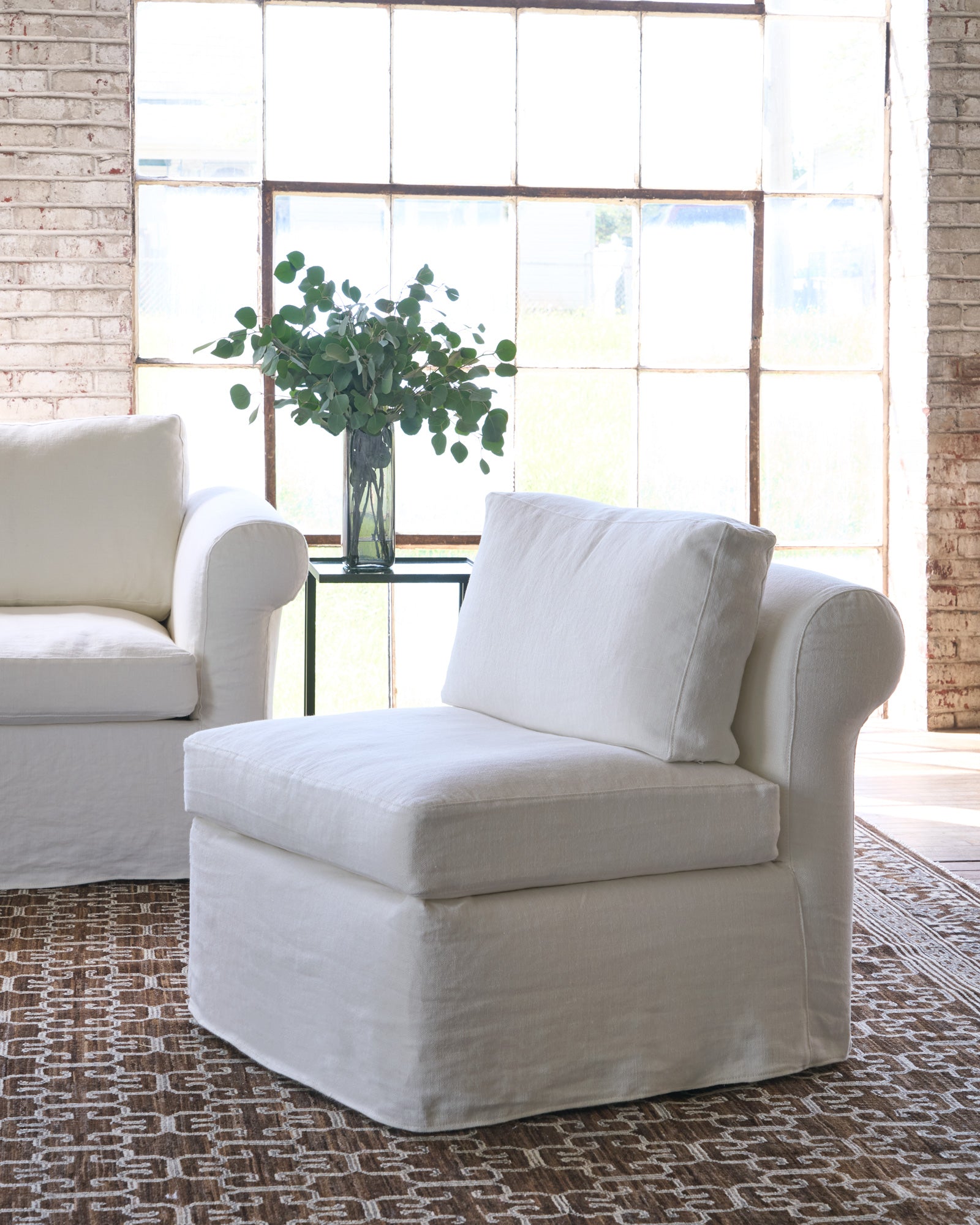  White chair on a brown rug next to a white sofa and side table with a plant on top. Photographed in Brevard Ivory. 