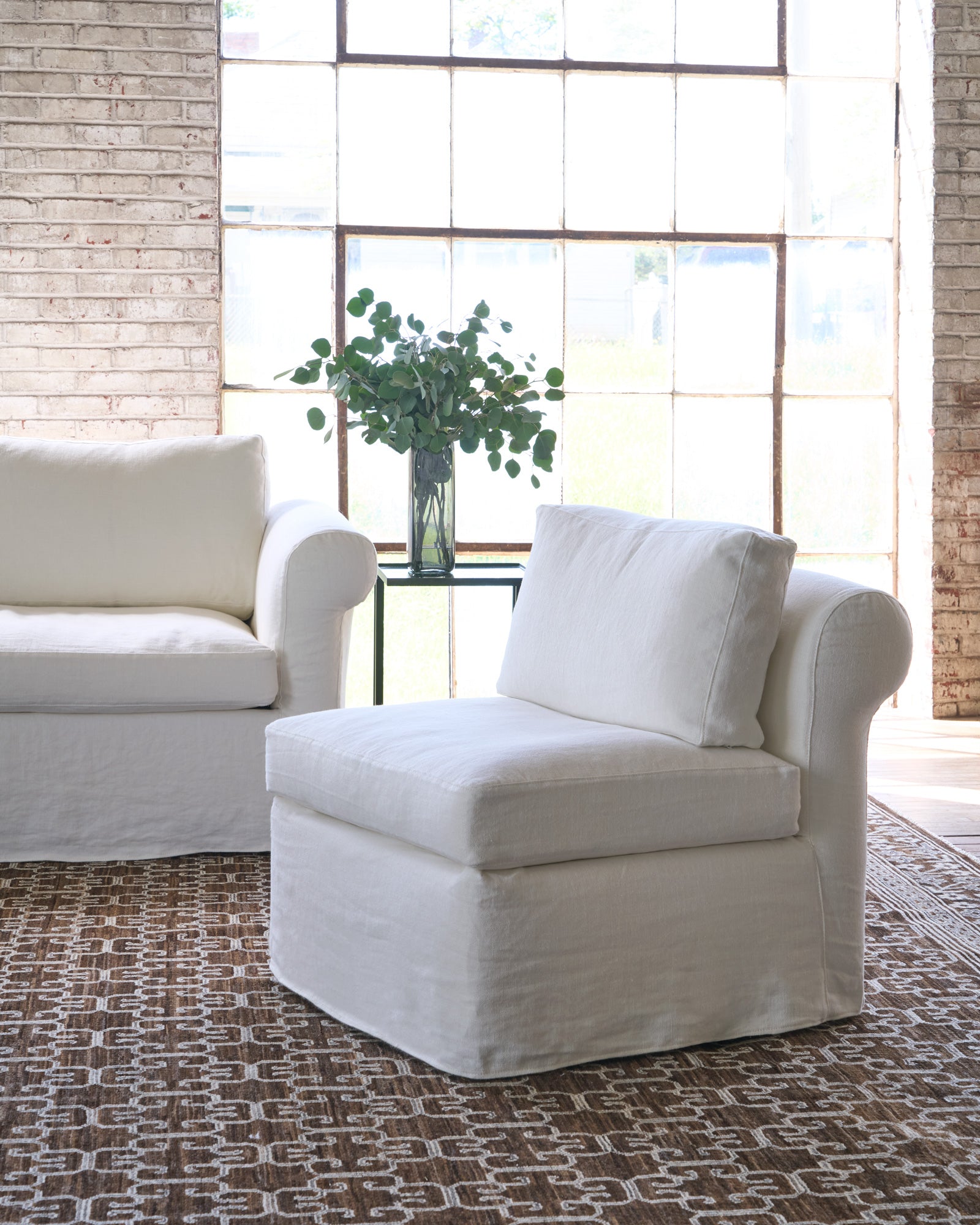  White chair on a brown rug next to a white sofa and side table with a plant on top. Photographed in Brevard Ivory. 