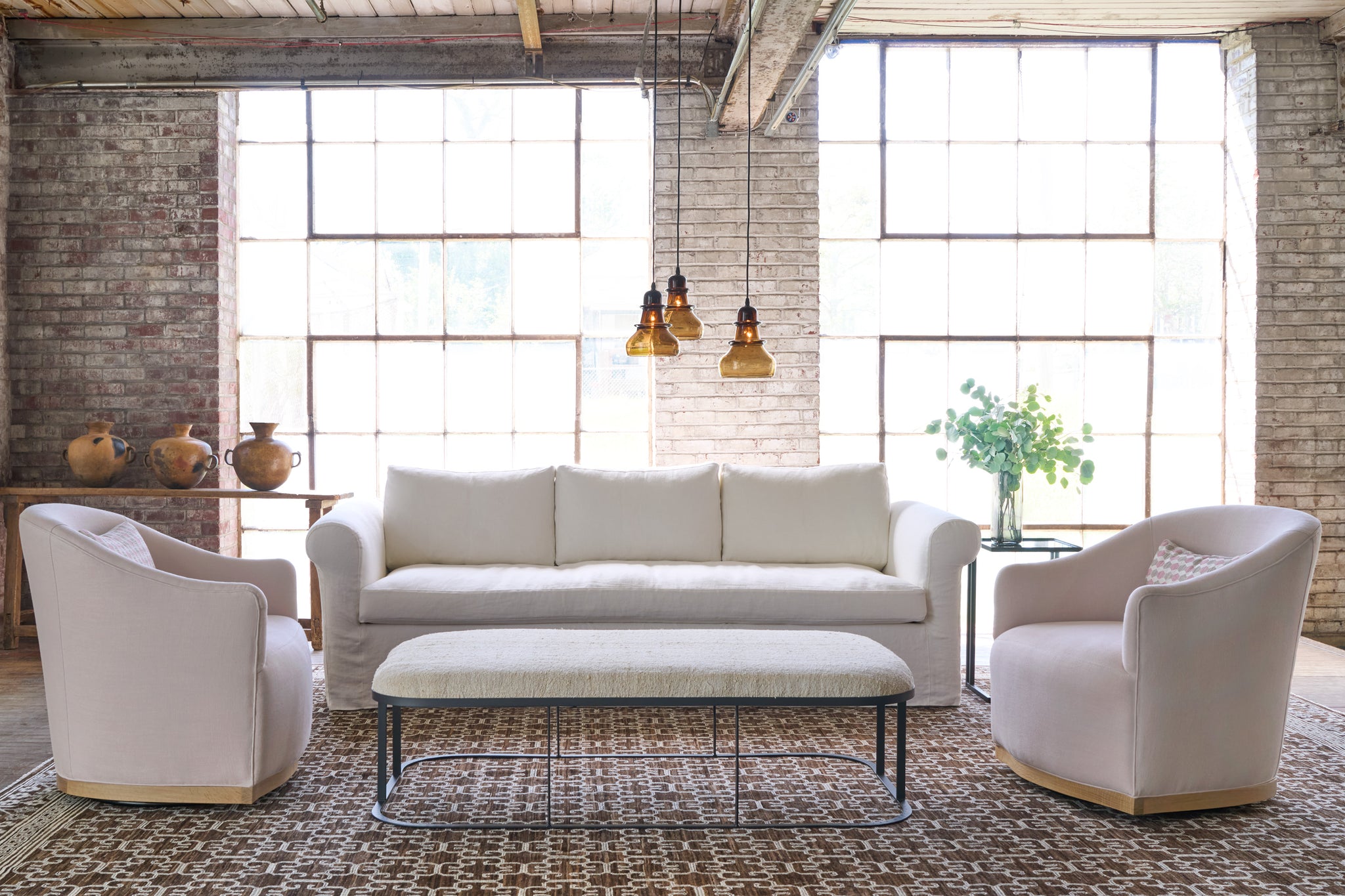  White sofa in a showroom with 2 pink chairs on the side. 3 brown glass pendants above an ottoman. Photographed in Brevard Rose. 