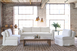  White chair son a brown rug next to a white sofa and side table with 3 pots on top. 2 large windows in the background and a wood coffee table in front. Photographed in Brevard Ivory. 