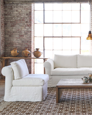  White chair on a brown rug next to a white sofa and side table with 3 pots on top. Photographed in Brevard Ivory. 