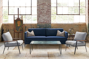  Blue sofa in a showroom with a coffee table with a mirro top. A chair on each side and a painting on the left side and s pot on the right. Photographed in Lester Ink. 