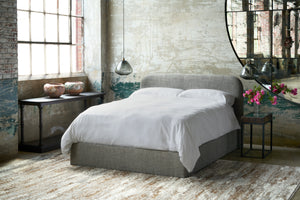  Bed in a showroom by a large window with smoke glass lights on the side. Wood console under the window, side table with pink flowers in front of the mirror. Photographed in Raven Peppercorn. 