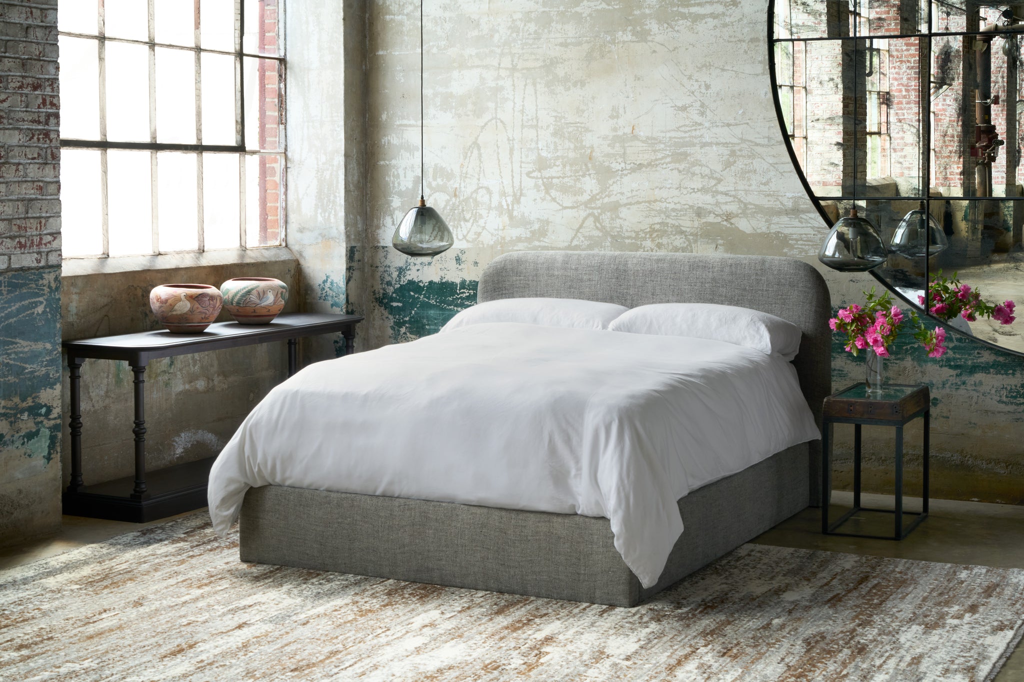  Bed in a showroom by a large window with smoke glass lights on the side. Wood console under the window, side table with pink flowers in front of the mirror. Photographed in Raven Peppercorn. 