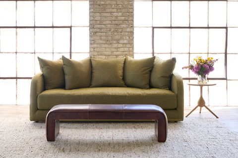 Green Sofa in front of 2 windows with a brown leather bench. Photographed in Spur Chocolate.