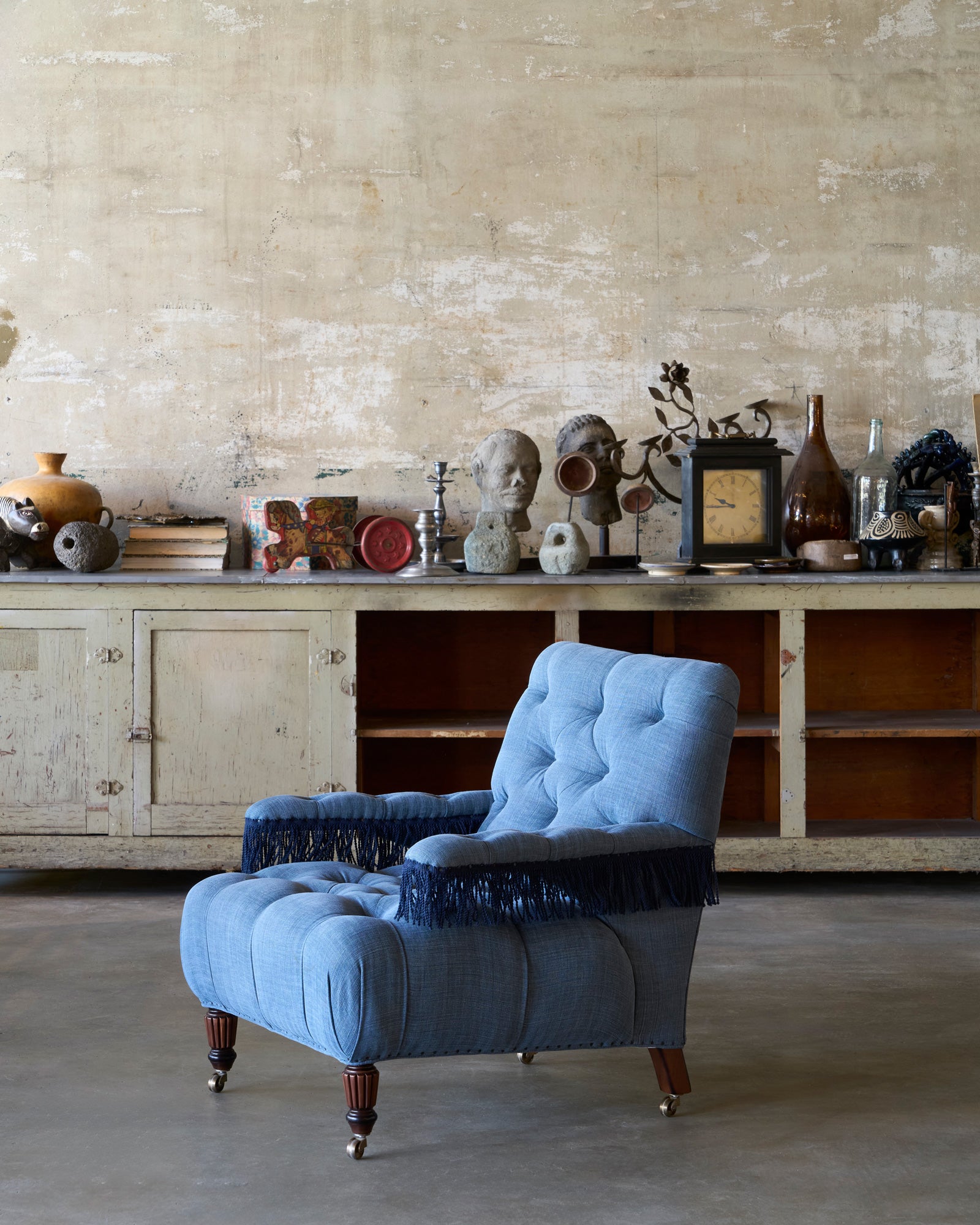  A blue tufted chair with fringes in front of a a console with various objects on top. Photographed in Rye Midnight Blue. 