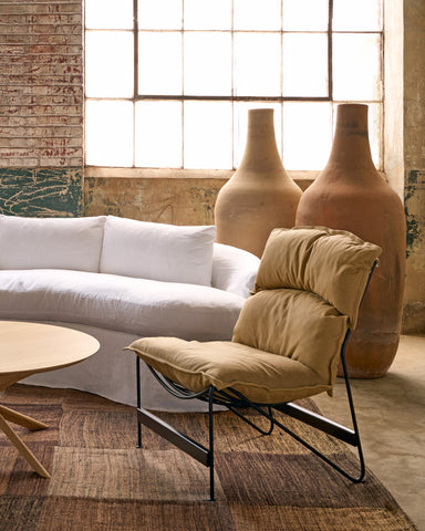 A modern lounge chair with a beige cushioned seat and backrest, supported by a black metal frame, positioned in a rustic living room next to a white sofa and large terracotta vases. Photographed in Bracco Tan.